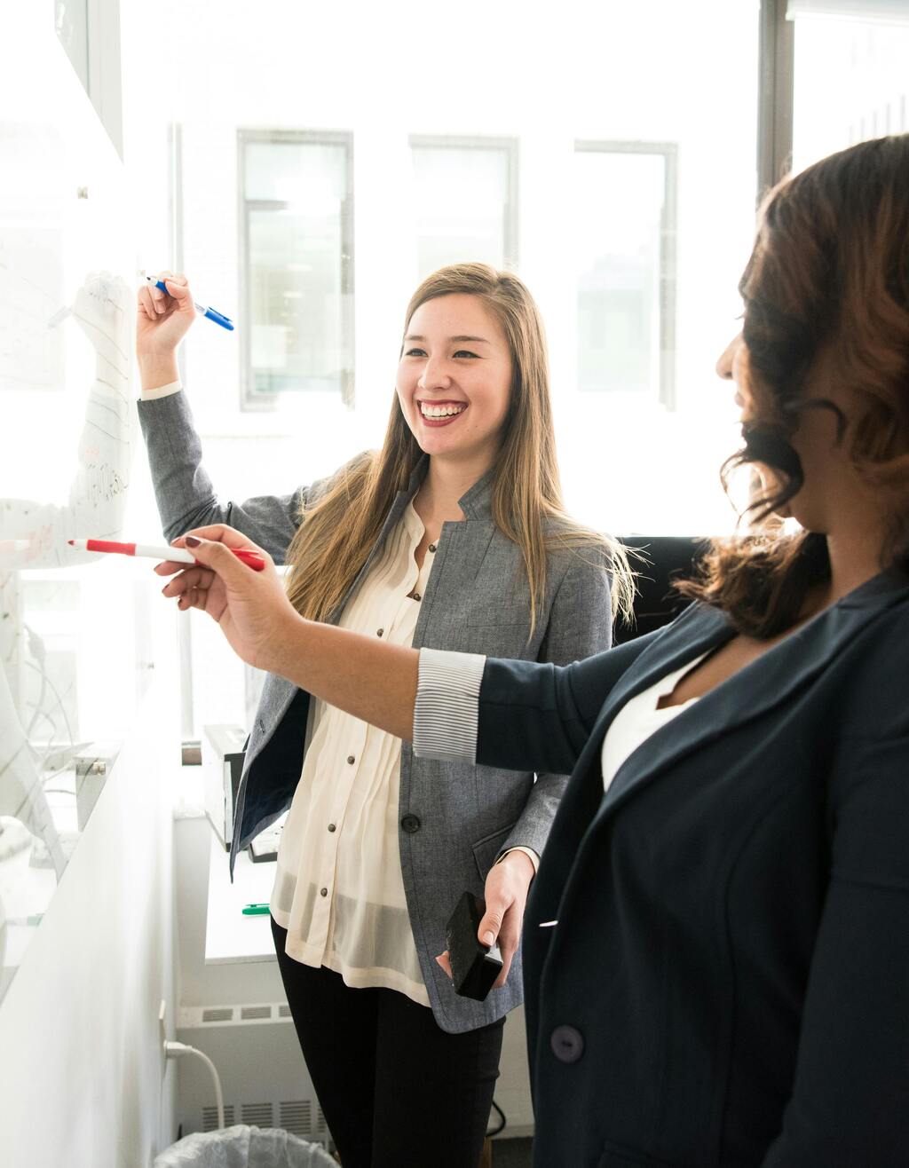 Two professionally-dressed women converse and smile as they make notations on an office whiteboard.