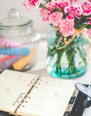 A daily planner sits open on a table with a vase of pink carnation flowers next to it.