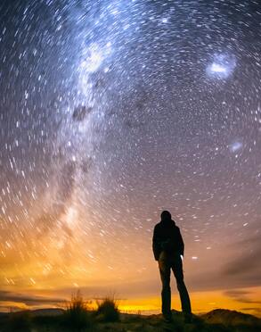 A silhouetted person stands on a ridge, gazing up into the night sky.  The stars are on a Timelapse photography, making them streaked across the sky.